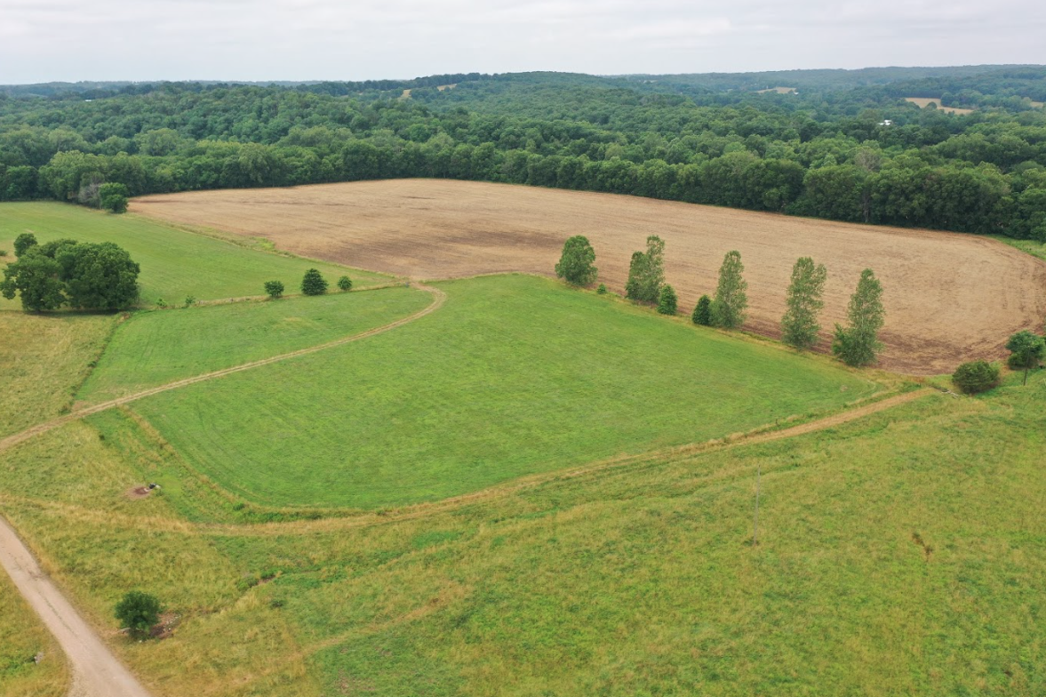 Aerial view of a rural landscape featuring a mix of green grassland and a brown field, bordered by trees and a dirt road, under a cloudy sky.