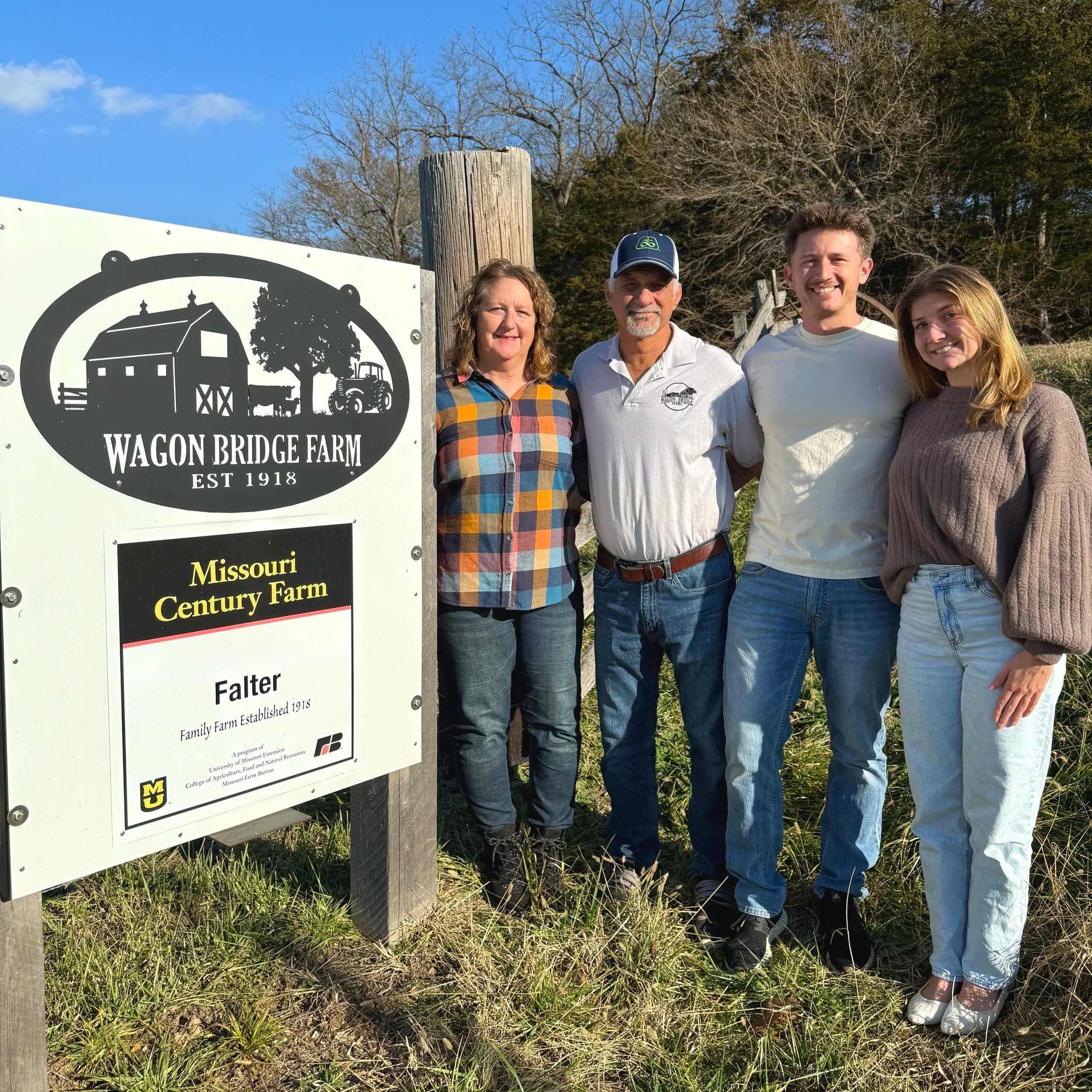 Family standing by the Wagon Bridge Farm sign in Missouri, celebrating its status as a Century Farm established in 1918. The group includes two adults and two younger individuals, all smiling and posing in front of the farm's logo and information.