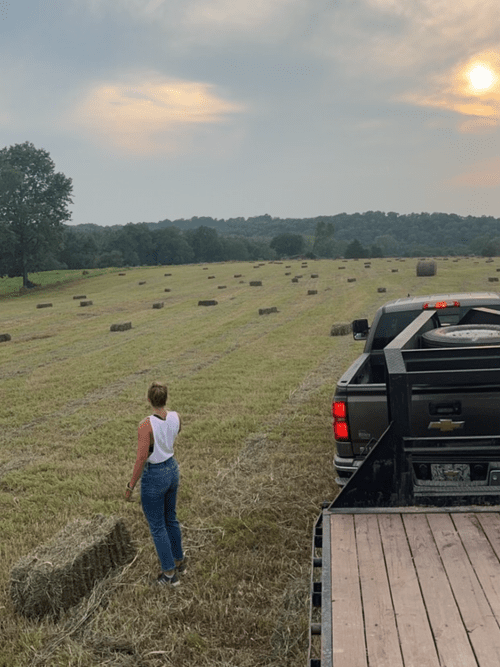 Woman in a white tank top and jeans standing in a hayfield with round bales scattered across the landscape, a pickup truck parked nearby under a hazy sky at sunset.
